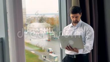 白天，一个商人在一台<strong>银色笔记本</strong>电脑上工作，站在一扇敞开的窗户附近。